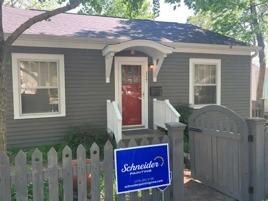 A gray cottage with a red front door and a Schneider Painting yard sign at the gate